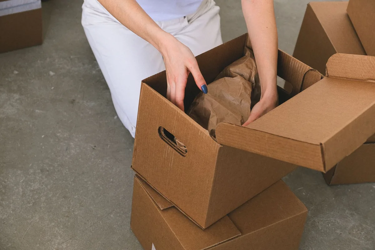 From above of crop anonymous relocating female packing wrapped items into carton box while preparing for moving to new apartment
