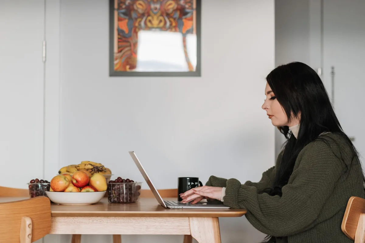 Side view of young female remote employee working on netbook at table with assorted fruits and hot drink at home
